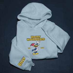 May include: Light blue hoodie featuring the cartoon character Woody Woodpecker. The design includes the text "WOODY WOODPECKER" above an image of the character, with "EST. 1940" below. The sleeve reads "BILLY" in yellow.