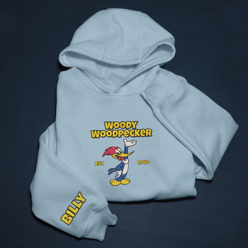 May include: Light blue hoodie featuring the cartoon character Woody Woodpecker. The design includes the text "WOODY WOODPECKER" above an image of the character, with "EST. 1940" below. The sleeve reads "BILLY" in yellow.