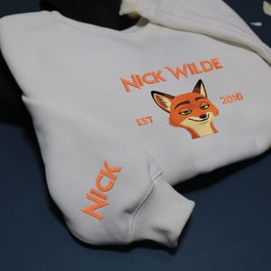 May include: White sweatshirt featuring orange embroidery. The design includes the text "NICK WILDE EST 2016" and a fox face on the chest. The sleeve displays the name "NICK" in orange. A casual, comfortable garment.