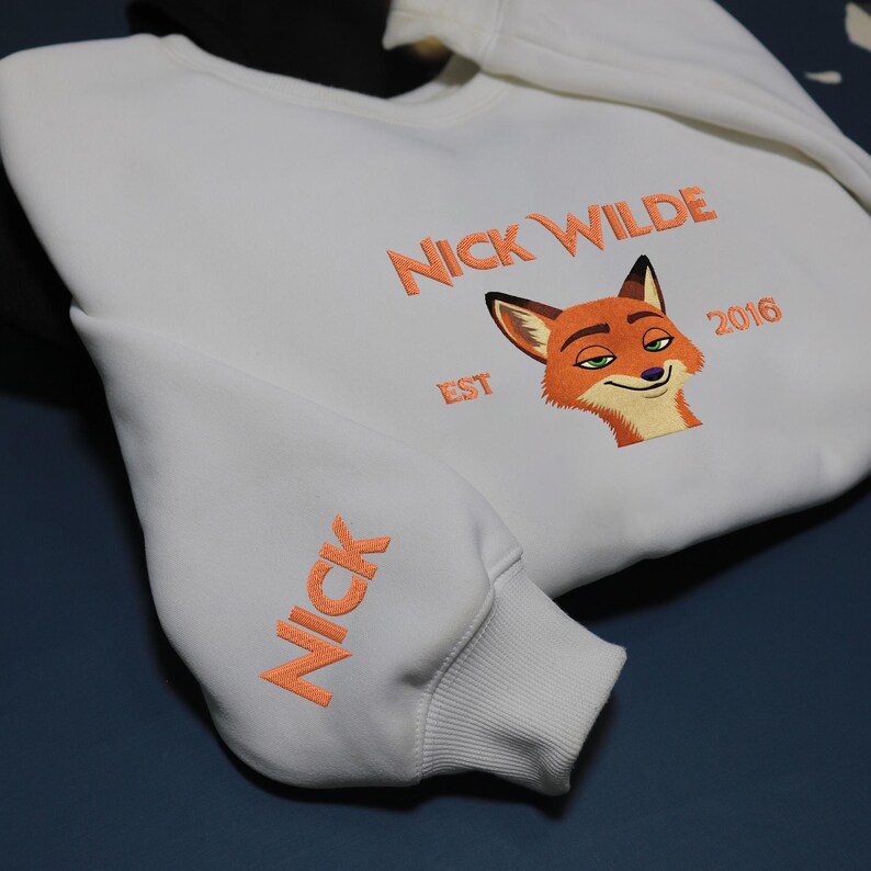 May include: White sweatshirt featuring orange embroidery. The design includes the text "NICK WILDE EST 2016" and a fox face on the chest. The sleeve displays the name "NICK" in orange. A casual, comfortable garment.