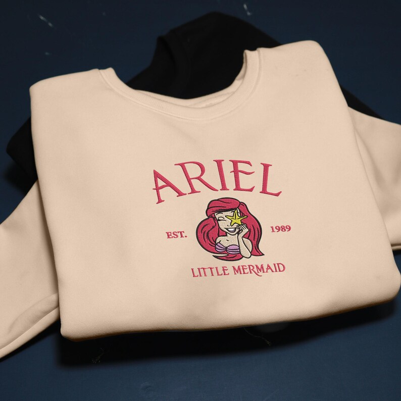 May include: A cream-coloured sweatshirt with the word "ARIEL" in pink script, and an illustration of Ariel from The Little Mermaid. The design includes the text "EST. 1989 LITTLE MERMAID". A black sweatshirt is visible underneath.
