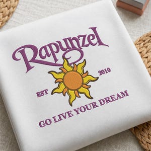 Può includere: Una felpa bianca con testo ricamato in viola che recita Rapunzel, EST 2010 e GO LIVE YOUR DREAM. Al centro, tra il testo, si trova un sole ricamato in giallo e arancione con raggi appuntiti a forma di fiamma e un centro circolare.