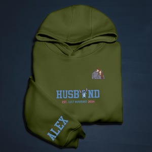 Puede incluir: Sudadera con capucha verde oliva con la palabra "HUSBAND" en azul, un personaje de dibujos animados y el texto "EST. JUST MARRIED 2024". La manga muestra el nombre "ALEX" en azul. Una pareja est&aacute; bordada en el pecho.