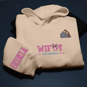 Puede incluir: Sudadera con capucha color crema con el nombre "SONIA" en letras rosas en la manga. La parte delantera presenta la palabra "WIFEY" con un personaje de dibujos animados y el texto "EST. JUST MARRIED 2024". Tambi&eacute;n hay una peque&ntilde;a pareja bordada.