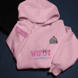 Puede incluir: Una sudadera con capucha rosa claro con la palabra "SONIA" en la manga izquierda. La sudadera presenta una pareja bordada y la palabra "WIFEY" con un personaje de dibujos animados. Tambi&eacute;n se ve el texto "1ST. JUST MARRIED 2024".