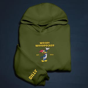 May include: Olive green hoodie featuring the cartoon character Woody Woodpecker. The hoodie has the text "WOODY WOODPECKER" in yellow, with the character illustration and the year "1940". The sleeve reads "BILLY" in yellow.