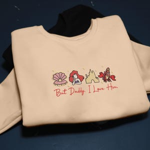 Pode incluir: Um moletom creme com um gr&aacute;fico com uma concha, uma sereia ruiva, um castelo e um caranguejo vermelho. O texto "But Daddy, I Love Him" est&aacute; abaixo do gr&aacute;fico. Um moletom preto &eacute; vis&iacute;vel no fundo.