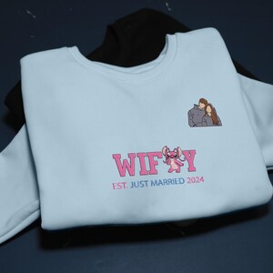 Puede incluir: Sudadera azul claro con la palabra "WIFEY" en rosa, un personaje de dibujos animados y el texto "EST. JUST MARRIED 2024". Una peque&ntilde;a ilustraci&oacute;n de una pareja est&aacute; en la parte superior derecha. Una sudadera negra es parcialmente visible.