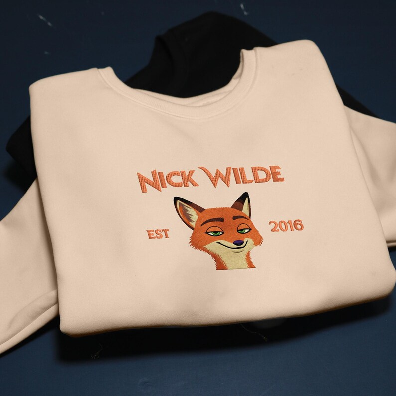 May include: Cream-coloured sweatshirt with the text "Nick Wilde" above a cartoon fox face. The text "EST 2016" is below the text and fox graphic. A black sweatshirt is visible underneath.