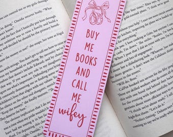 May include: A pink bookmark with the text "BUY ME BOOKS AND CALL ME wifey" in red. The bookmark has a decorative border and a bow and rings graphic at the top, and is placed on an open book.