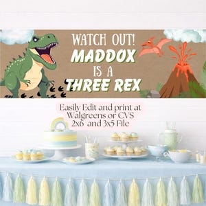 May include: A rectangular banner featuring a green Tyrannosaurus Rex, a flying pterodactyl, and an erupting volcano on a brown background. The text reads: WATCH OUT! MADDOX IS A THREE REX. The design includes white clouds and green foliage elements.