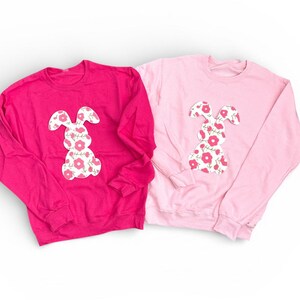 May include: Two long-sleeved sweatshirts, one in a vibrant pink and the other a pale pink. Each has a white bunny silhouette with a floral design. The bunny motif is positioned centrally on the front of the shirts, giving a cheerful appearance.