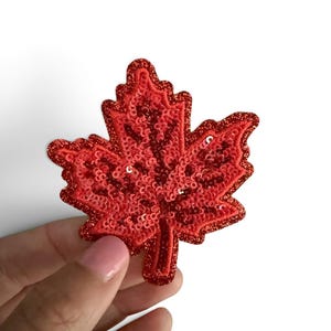 May include: A red maple leaf patch featuring a dense layer of shimmering sequins and glittery thread detailing. The embroidered design captures the iconic five-pointed shape of a maple leaf with a textured surface and a defined central stem.