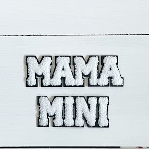 May include: White felt letters spelling "MAMA MINI" on a white wooden background.