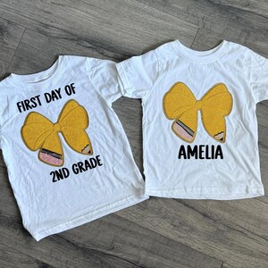 May include: Two white t-shirts with a yellow bow design. One shirt says "FIRST DAY OF 2ND GRADE" and the other says "AMELIA" in black letters. The bow design has a pink and black pencil detail.