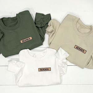 May include: Three sweatshirts in different colors, green, beige, and white, with the word "MAMA" printed on each in a brown rectangle.