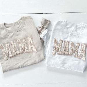 May include: Two beige and white long-sleeved shirts with the word "MAMA" in floral print letters.