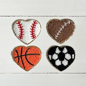 May include: Four felt heart-shaped patches with different sports designs. The patches are white, brown, orange, and black with white. The patches are outlined in gold.