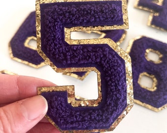 Puede incluir: Número 5 de fieltro morado con ribete de purpurina dorada. El número es grueso y tiene una textura ligeramente en relieve.