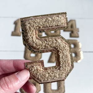 May include: A gold glitter and fuzzy felt number five patch. The patch has a gold glitter outline and a fuzzy brown felt interior.