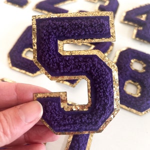 May include: Purple felt number 5 with gold glitter trim. The number is thick and has a slightly raised texture.