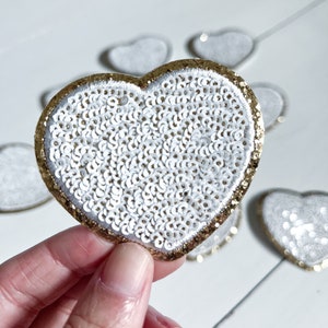 May include: A white heart-shaped patch with a gold glitter border, covered in white sequins.
