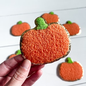 May include: Five orange felt pumpkins with gold glitter trim and green felt stems. The pumpkins are arranged in a semi-circle on a white surface.