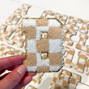 May include: A beige and white checkered letter B with gold glitter trim. The letter is made of a fluffy material and is surrounded by other letters in the same style.