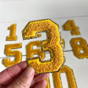 May include: A set of yellow felt and gold glitter iron-on patches in the shape of numbers. The numbers are 1, 3, 4, 5, 6, 8, and 10.