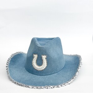 May include: A light blue denim cowboy hat with a silver sequin trim and a white horseshoe design. The horseshoe is embellished with pearls and rhinestones. The hat has a classic cowboy hat shape.