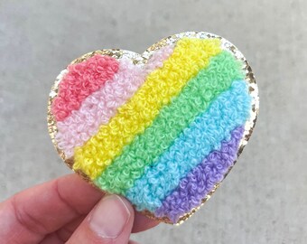May include: A heart-shaped patch with a rainbow design. The patch features a gold glitter border and a textured, looped fabric in red, pink, yellow, green, blue, and purple. The patch is held by a hand.