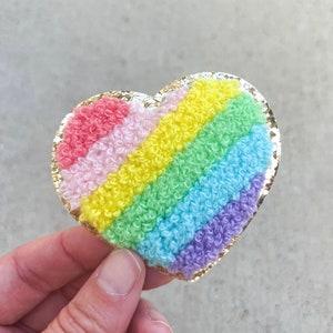 May include: A heart-shaped patch with a rainbow design. The patch features a gold glitter border and a textured, looped fabric in red, pink, yellow, green, blue, and purple. The patch is held by a hand.