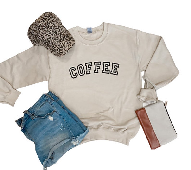 K&ouml;nnte beinhalten: Ein cremefarbenes Sweatshirt mit dem Wort "COFFEE" in schwarzen Buchstaben. Dazu geh&ouml;ren Jeansshorts, ein Leopardenmuster-Hut und eine zweifarbige Clutch. Die Artikel sind auf wei&szlig;em Hintergrund angeordnet.