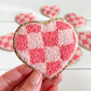 May include: A pink and white checkered heart-shaped patch with a gold glitter border. The patch is made of a soft, fuzzy material.
