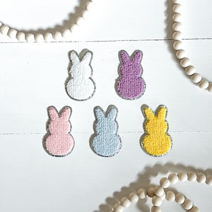May include: Five fuzzy bunny shaped patches in different colours. The patches are white, pink, purple, blue, and yellow. Each patch has a silver glitter outline.
