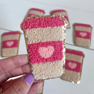May include: A set of five pink and beige coffee cup patches with gold glitter trim and a pink heart design. The patches are made of fuzzy yarn and are perfect for adding a touch of whimsy to any project.