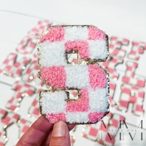 May include: Pink and white checkered letter S with gold glitter trim. The letter is made of fluffy fabric and is a patch or applique.