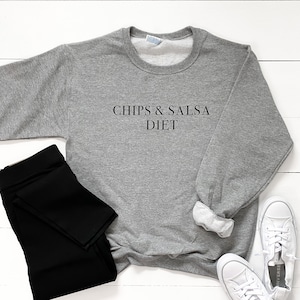 May include: Grey sweatshirt with the text "Chips & Salsa Diet" printed on the front. The sweatshirt is laid out on a white wooden surface with black leggings and white trainers.