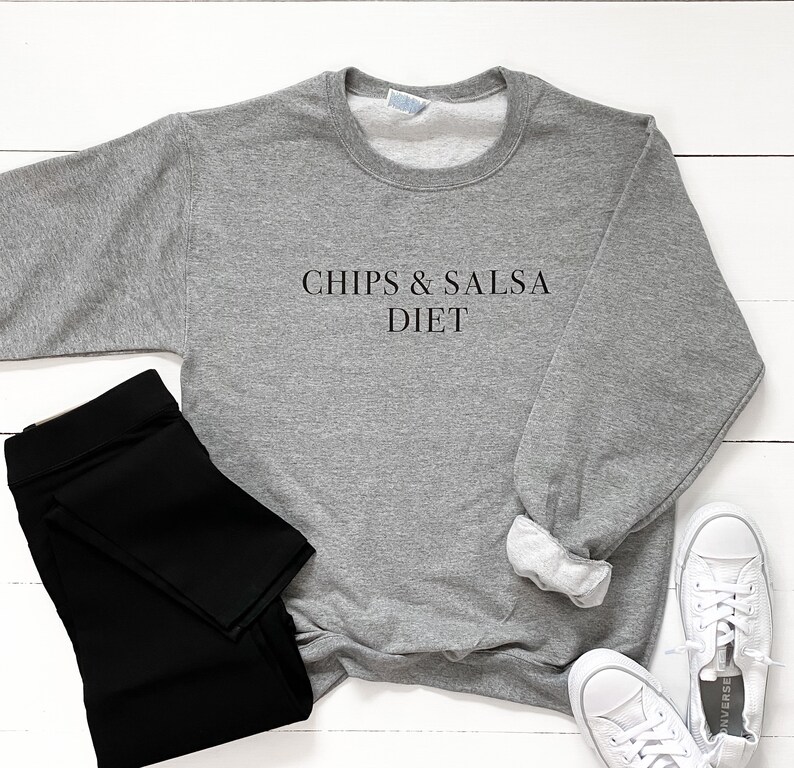 May include: Grey sweatshirt with the text "Chips & Salsa Diet" printed on the front. The sweatshirt is laid out on a white wooden surface with black leggings and white trainers.