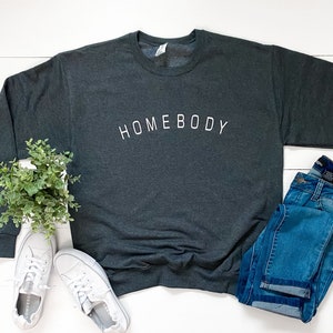 Puede incluir: Una sudadera gris oscuro con la palabra "HOMEBODY" impresa en blanco en la parte delantera. La sudadera est&aacute; colocada sobre una superficie de madera blanca con un par de zapatillas blancas, una planta en maceta y un par de pantalones vaqueros azules.