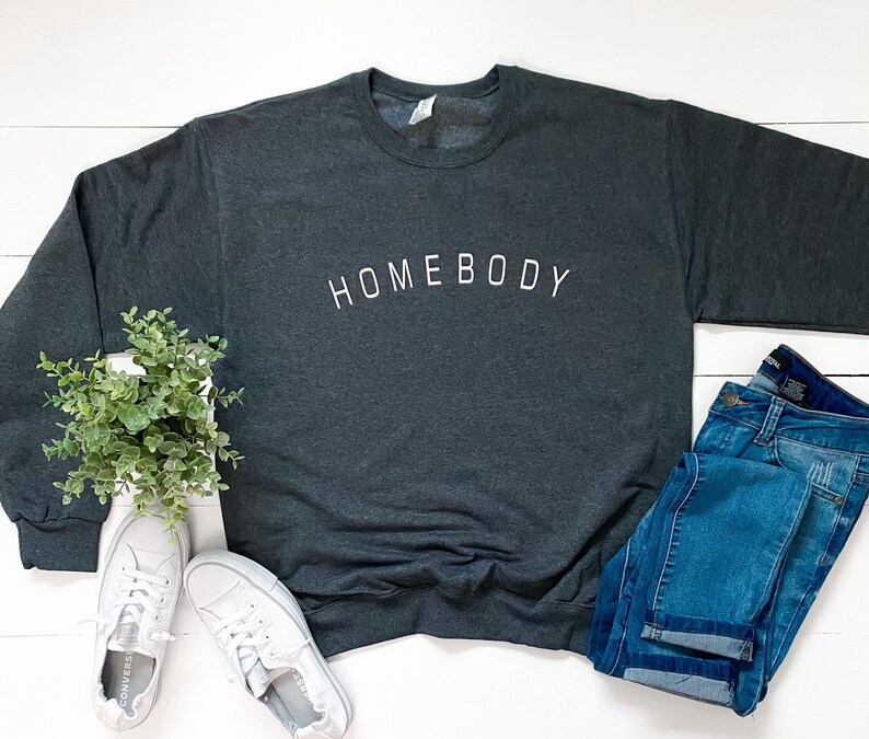 Puede incluir: Una sudadera gris oscuro con la palabra "HOMEBODY" impresa en blanco en la parte delantera. La sudadera est&aacute; colocada sobre una superficie de madera blanca con un par de zapatillas blancas, una planta en maceta y un par de pantalones vaqueros azules.