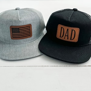 May include: Two trucker hats with brown leather patches. One hat is gray with a US flag patch and the other is black with a "DAD" patch.