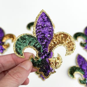May include: A sequined fleur-de-lis in purple, green, and gold, with a gold glitter border. The emblem is held in a hand, with other similar emblems in the background. The design is reminiscent of Mardi Gras.
