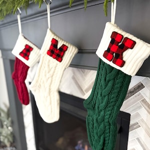 May include: Three Christmas stockings hanging on a fireplace mantel. The stockings are made of knitted fabric in red, white, and green. Each stocking has a red and black plaid letter on the front. The letters are "I", "M", and "K".