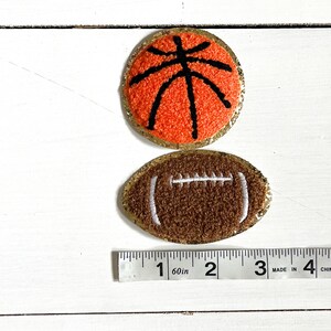 May include: Two felt sports ball patches with gold glitter trim. A brown football with white stitching and an orange basketball with black stitching. A ruler is shown for size reference.