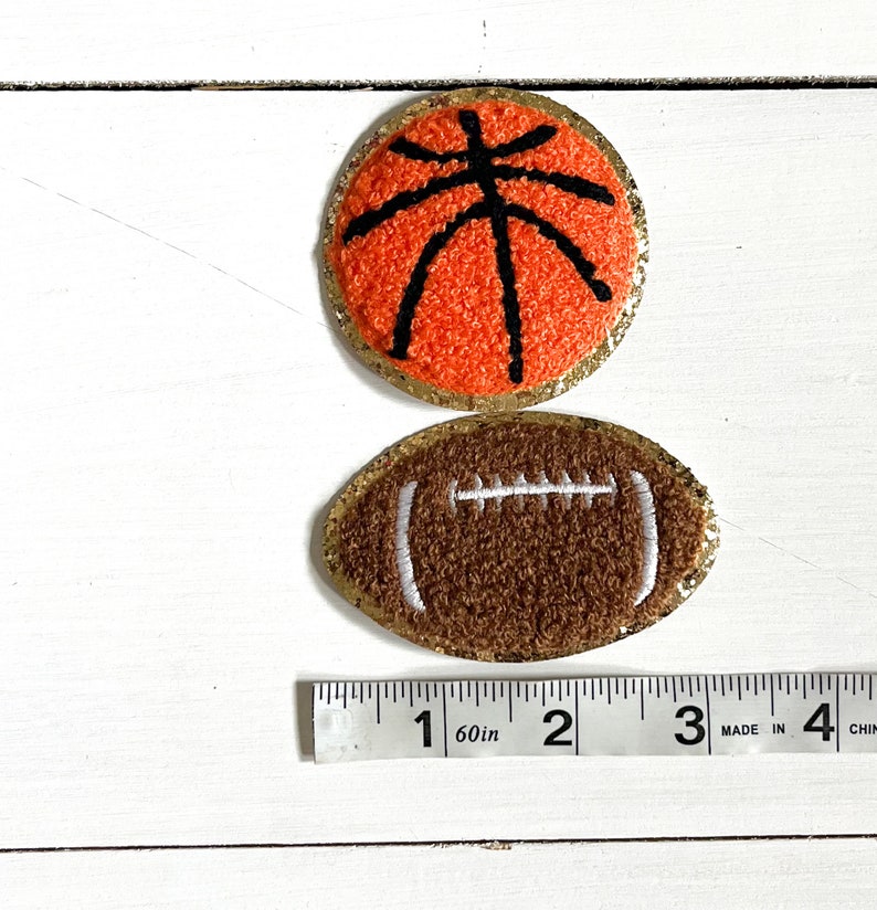 May include: Two felt sports ball patches with gold glitter trim. A brown football with white stitching and an orange basketball with black stitching. A ruler is shown for size reference.