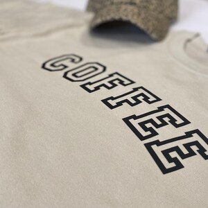K&ouml;nnte beinhalten: Beiges T-Shirt mit dem Wort &bdquo;COFFEE&ldquo; in fetter, schwarzer, collegeartiger Schrift vertikal aufgedruckt. Ein Hut mit Tweedmuster ist im Hintergrund teilweise zu sehen. Das Shirt besteht aus einem weichen Material.