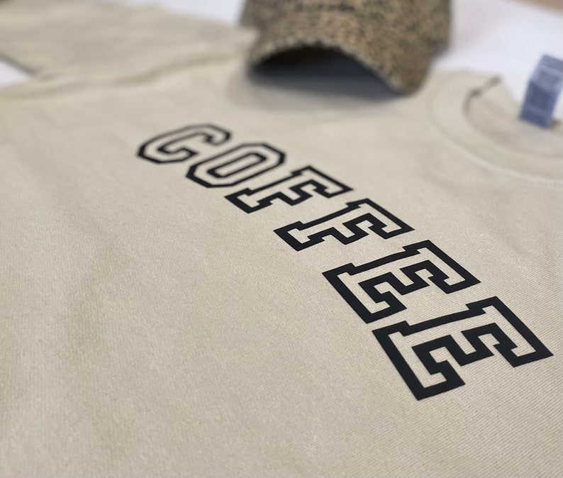 K&ouml;nnte beinhalten: Beiges T-Shirt mit dem Wort &bdquo;COFFEE&ldquo; in fetter, schwarzer, collegeartiger Schrift vertikal aufgedruckt. Ein Hut mit Tweedmuster ist im Hintergrund teilweise zu sehen. Das Shirt besteht aus einem weichen Material.
