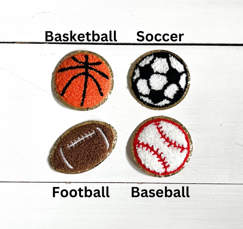 May include: Four embroidered felt patches depicting different sports balls: a basketball, soccer ball, football, and baseball. Each patch has a gold trim and is labeled with the sport's name.