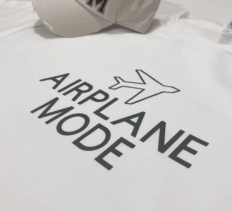 Puede incluir: Camiseta blanca con texto negro que dice "MODO AVI&Oacute;N" y un peque&ntilde;o gr&aacute;fico de avi&oacute;n.